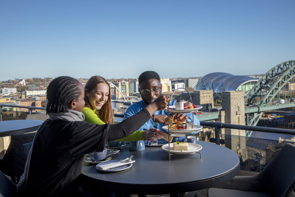 Three people enjoying afternoon tea on a rooftop with the River Tyne in the background