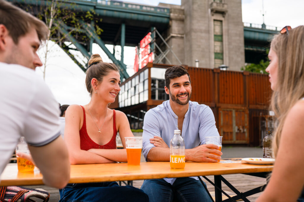 Four people enjoying drinks outside under the Tyne Bridge at a table.