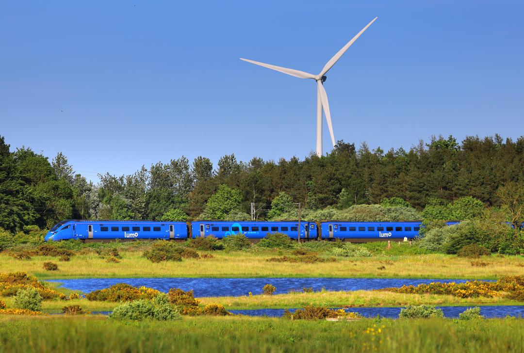 A lumo train going through the countryside, surrounded by greenery and with a wind turbine in the background