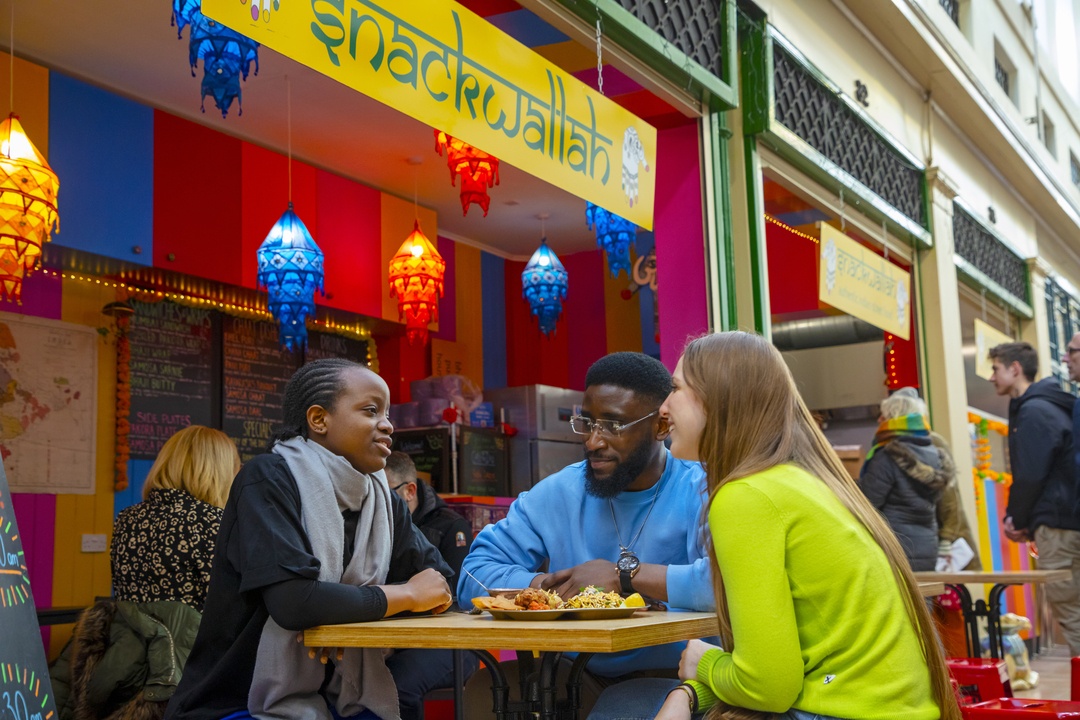 Three people sat at a table enjoying food at Snackwallah in Grainger Market. The stall is brightly multi-coloured.