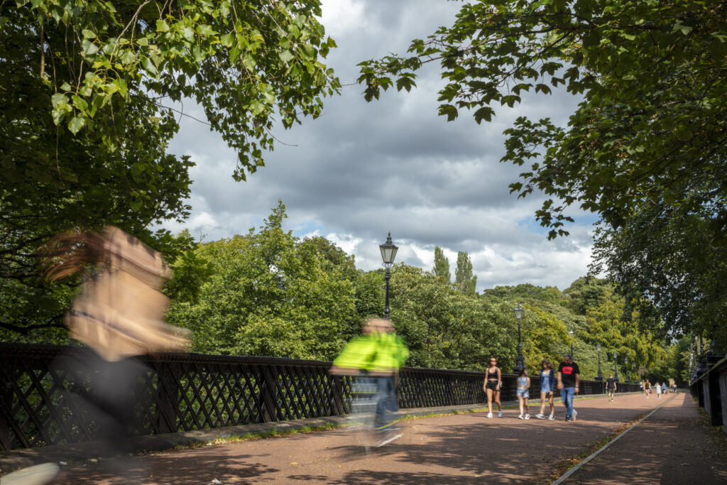 People walking, cycling and running across Armstrong Bridge in Jesmond Dene. The bridge is surrounded by leafy green trees.