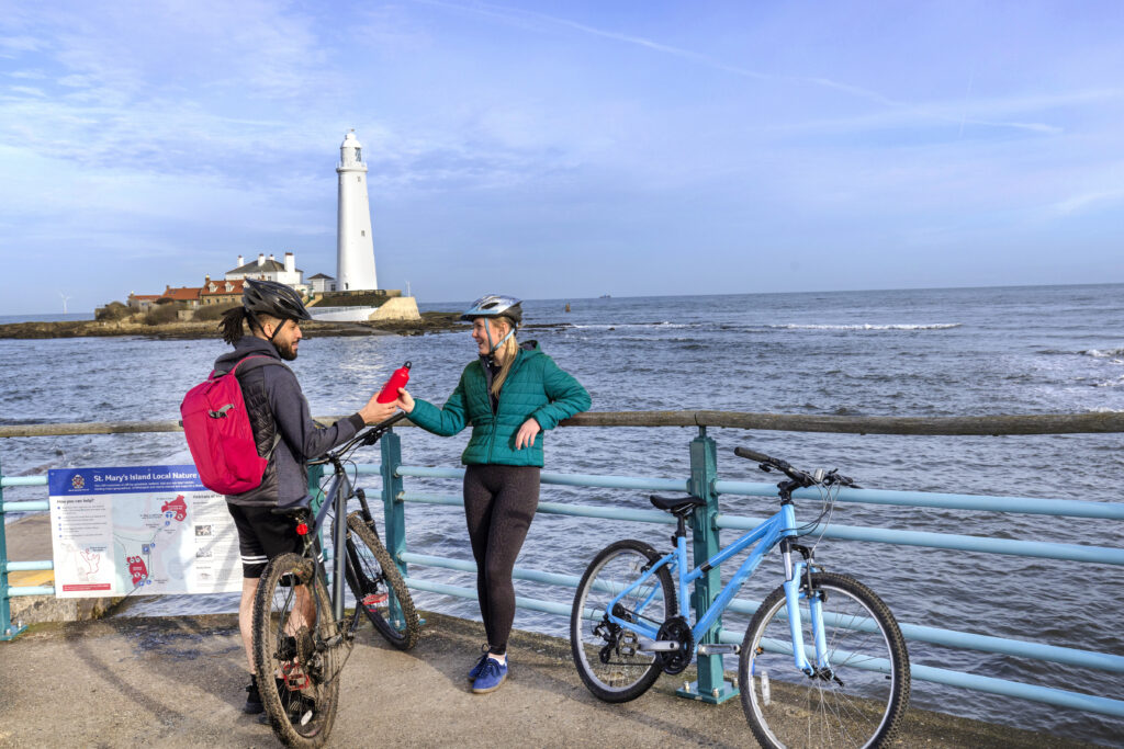 Two people stood at the barrier between the footpath and the sea with St Mary's Lighthouse in the background. They are both wearing helmets and with bicycles. One person hands the other a bottle of water.